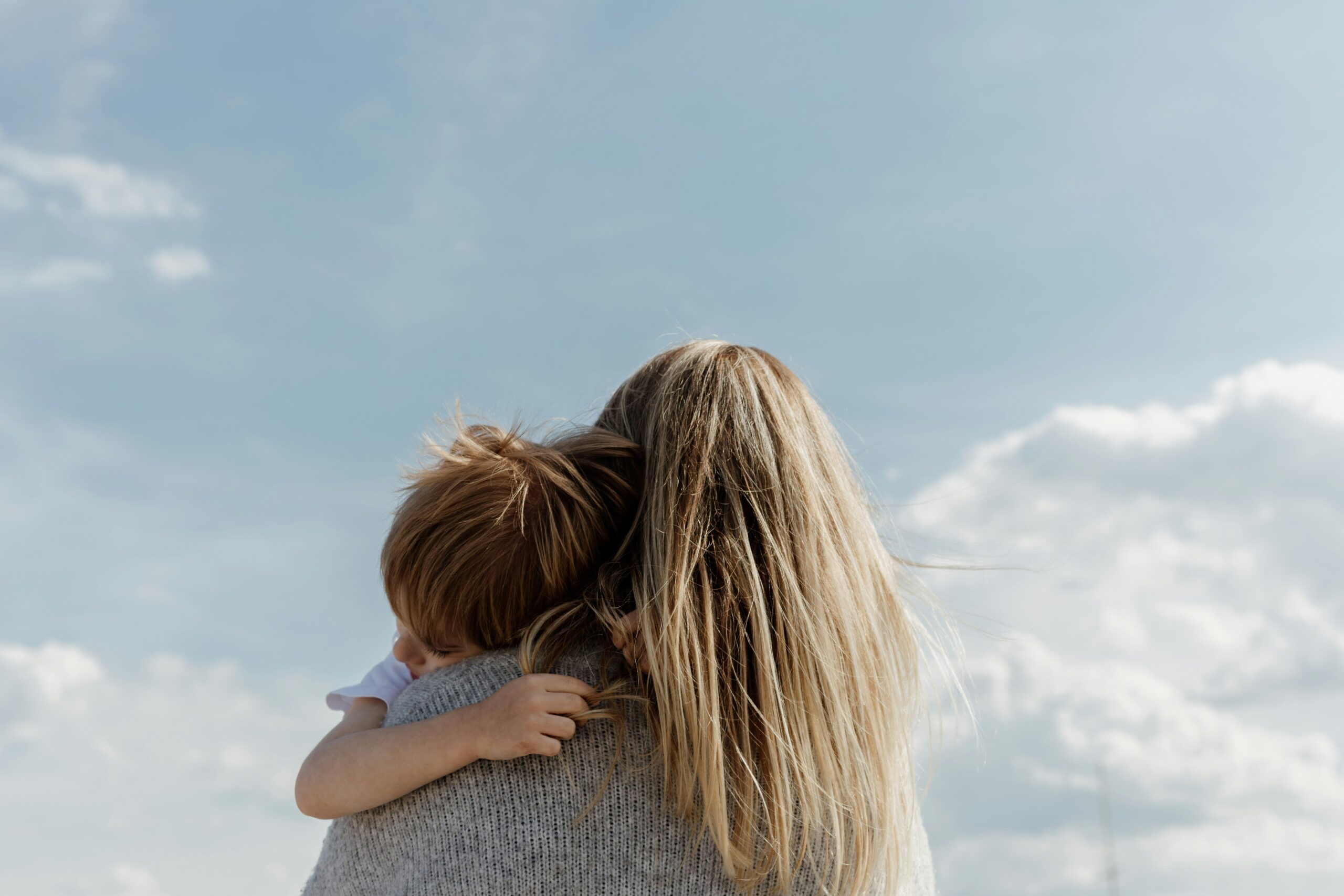 A woman with long blonde hair holds a child against a blue sky with clouds