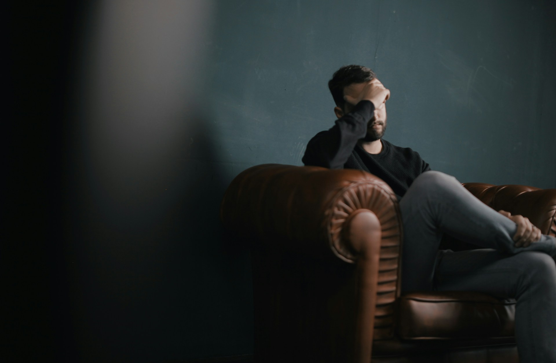man sitting on couch in dark room with face resting in hand.